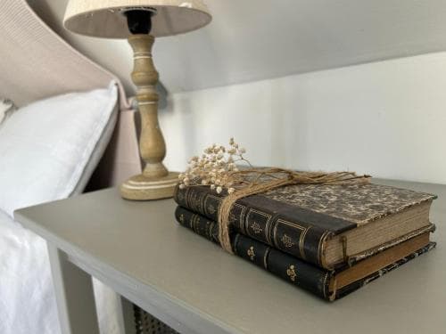 A cozy bedroom bedside table features a stack of vintage books tied with twine and baby's breath, alongside a rustic lamp, evoking a serene and nostalgic atmosphere ideal for a relaxing getaway.