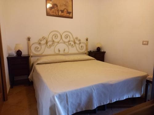 A cozy hotel room features a neatly made bed with an ornate white metal headboard, flanked by wooden nightstands with lamps, under a warm-toned wall adorned with a framed painting, ideal for travelers seeking comfort and simplicity.