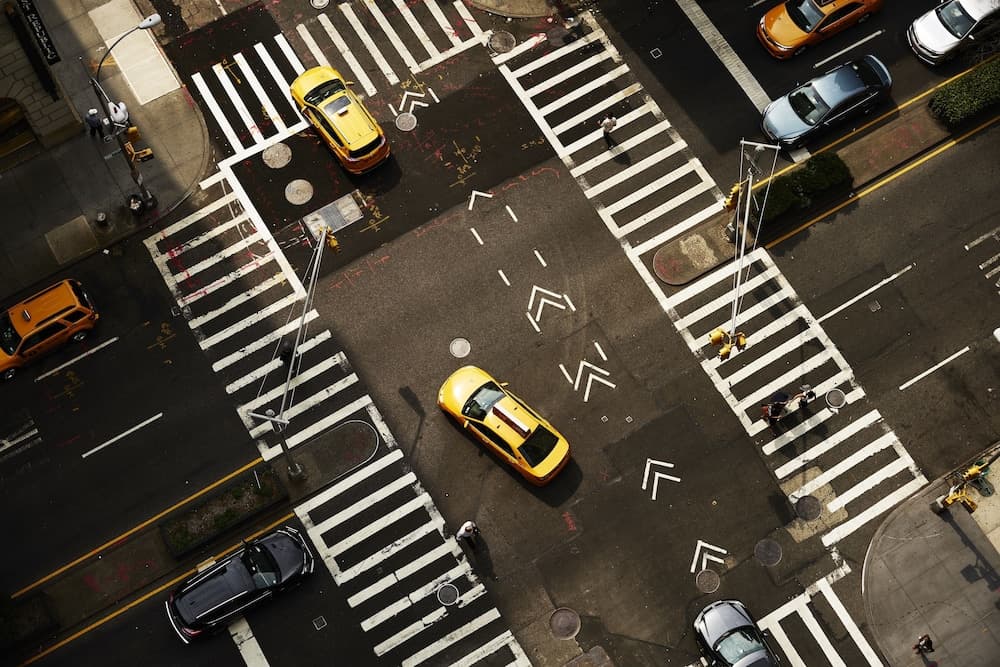 An aerial view captures the bustling urban energy of a city intersection filled with iconic yellow taxis, pedestrians crossing zebra-striped crosswalks, and a mix of vehicles, ideal for travelers exploring metropolitan life.