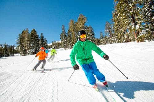 Skiers glide down a sunny, snow-covered slope surrounded by evergreen trees, capturing the joyful essence of a winter sports getaway in a picturesque mountain resort.