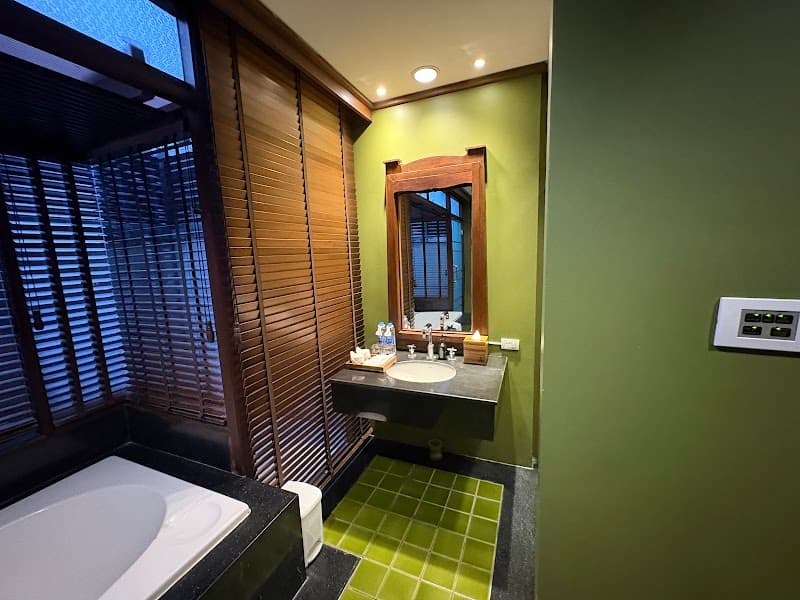 A serene bathroom at Nora Buri Resort & Spa in Koh Samui, Thailand, features a modern bathtub, green-tiled floor, wooden blinds, and a cozy ambiance ideal for travelers seeking relaxation.