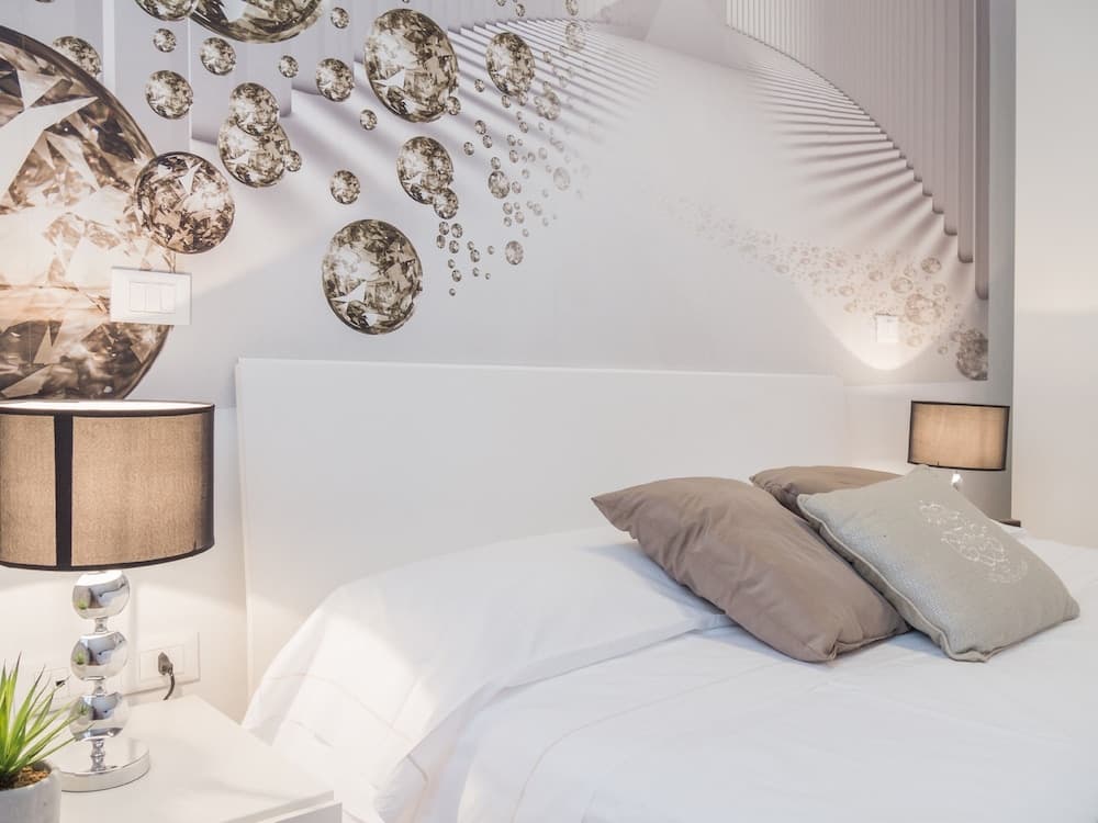 A serene bedroom features a bed adorned with neutral pillows against a wall decorated with sparkling diamond decals, complemented by elegant table lamps and a minimalist nightstand, ideal for travelers seeking a luxurious and tranquil retreat.