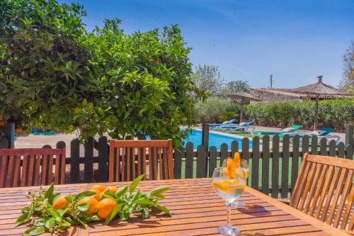 A serene outdoor dining area features a wooden table set with fresh oranges and a glass of water, surrounded by lush greenery and overlooking a tranquil poolside scene, ideal for a relaxing vacation retreat.