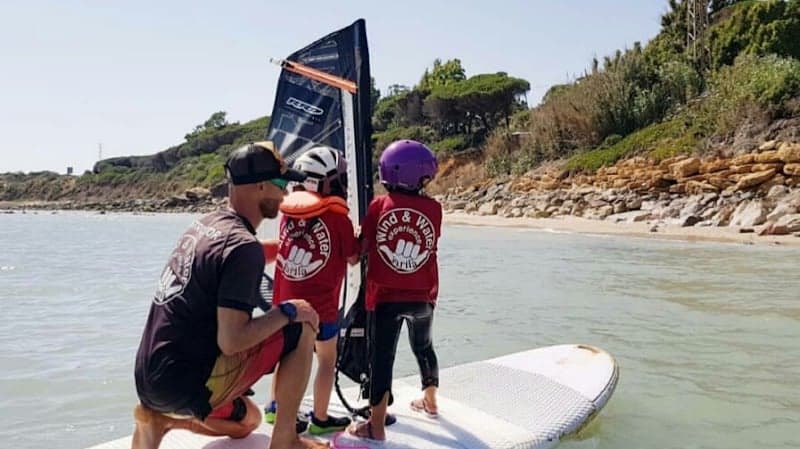 A sunny beach scene captures an instructor guiding two children in windsurfing gear on calm waters, ideal for family-friendly water sports adventures near lush green cliffs.