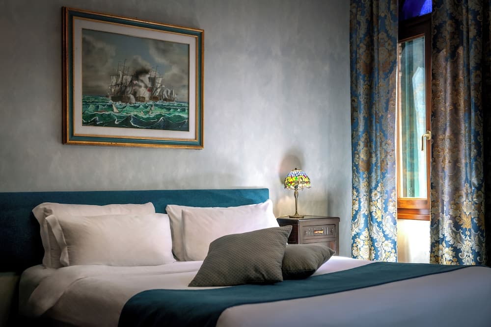 A cozy hotel room features a neatly made bed adorned with plush pillows, a vibrant painting of ships at sea above it, a stained-glass lamp on the bedside table, and elegant blue patterned curtains framing a window, ideal for travelers seeking comfort and charm.