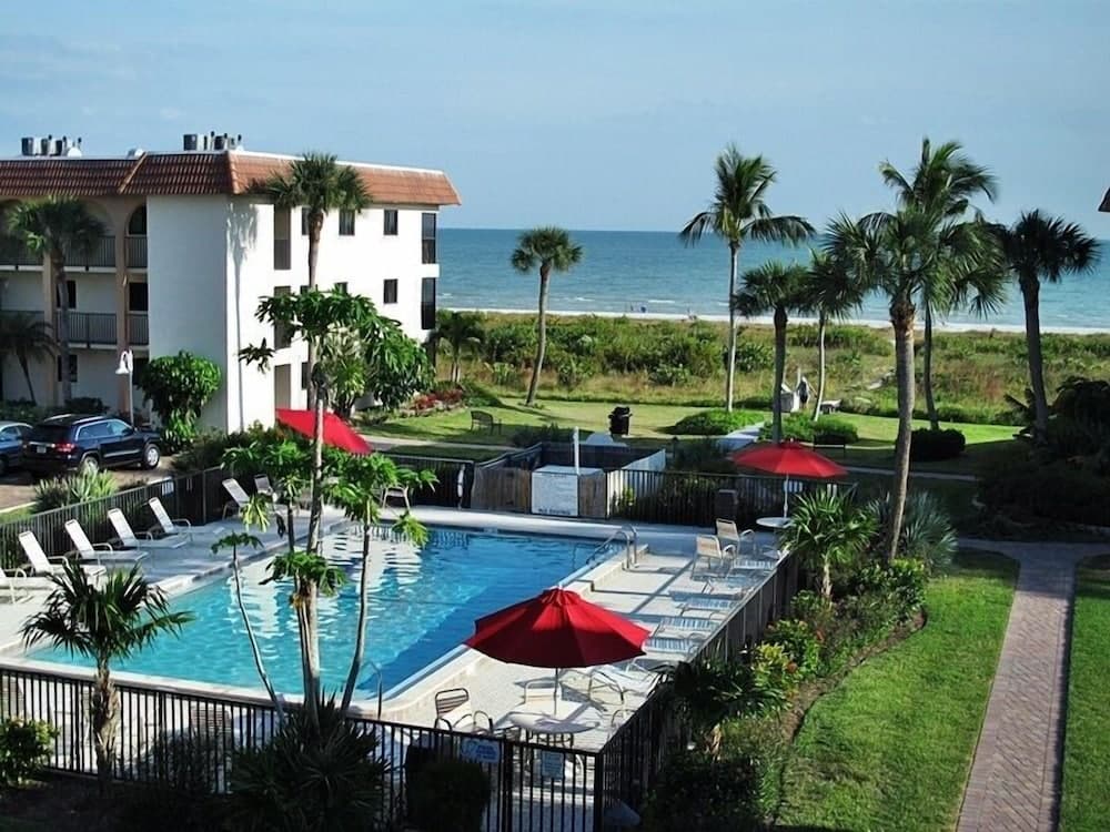 A serene coastal resort features a vibrant poolside area with lounge chairs and red umbrellas, surrounded by lush palm trees and overlooking the tranquil ocean, ideal for a relaxing beach vacation getaway.