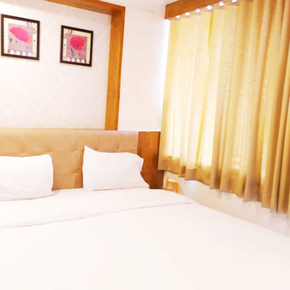 A cozy hotel room features a neatly made bed with white linens, framed floral artwork on the wall, and golden curtains, ideal for travelers seeking comfort and relaxation.