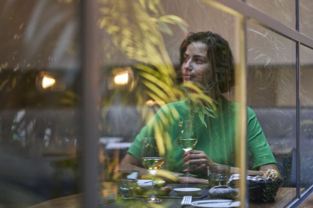 A serene dining scene captured through a window pane shows a woman enjoying wine amidst lush greenery, ideal for travelers seeking cozy, intimate restaurant experiences.