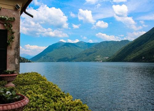 A serene lakeside view framed by lush greenery and distant mountains under a partly cloudy sky, ideal for tranquil retreats and scenic photography.