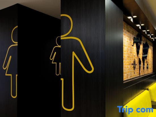 A modern restroom entrance features bold yellow gender symbols on black panels, complemented by a warmly lit brick wall adorned with artistic silhouettes, creating an inviting atmosphere ideal for travelers seeking stylish accommodations.