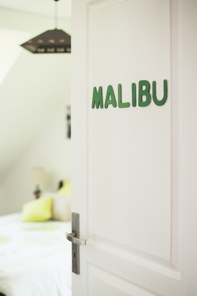 A cozy bedroom with a white door labeled "MALIBU" in green letters invites travelers seeking a serene retreat, featuring soft bedding and ambient lighting perfect for relaxation.