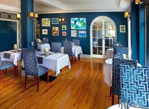 A cozy restaurant interior features elegant blue patterned chairs, white tablecloths on wooden floors, and vibrant artwork against deep blue walls, creating an inviting atmosphere perfect for a romantic dinner or special occasion.