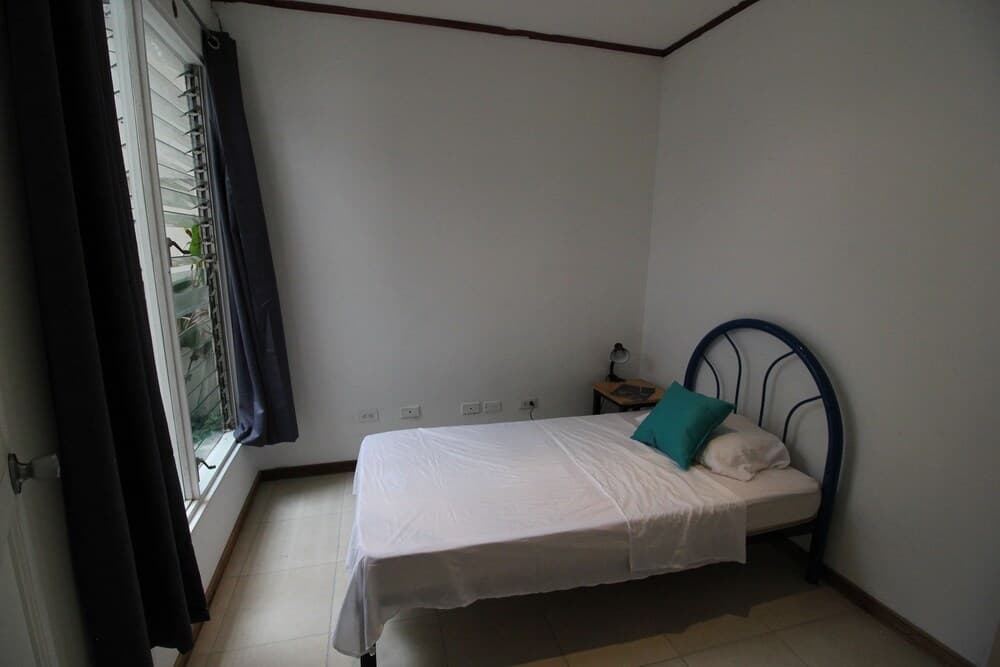A cozy minimalist bedroom with a single bed adorned with white linens and a teal pillow, featuring a small bedside table with a lamp, a window with dark curtains letting in soft light, ideal for budget travelers seeking simple accommodations.