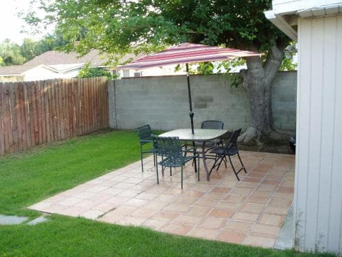 A cozy backyard patio features a tiled seating area with a table, chairs, and a striped umbrella under the shade of a large tree, ideal for relaxing or hosting small gatherings in a private outdoor space.