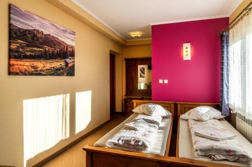 A cozy hotel room features two neatly made wooden beds under soft sunlight streaming through blinds, complemented by a vibrant pink accent wall and a scenic mountain artwork, ideal for travelers seeking comfort and relaxation.