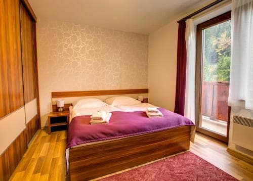 A cozy hotel room with a neatly made bed, wooden furnishings, and a large window offering a serene view of greenery, ideal for travelers seeking relaxation and comfort.
