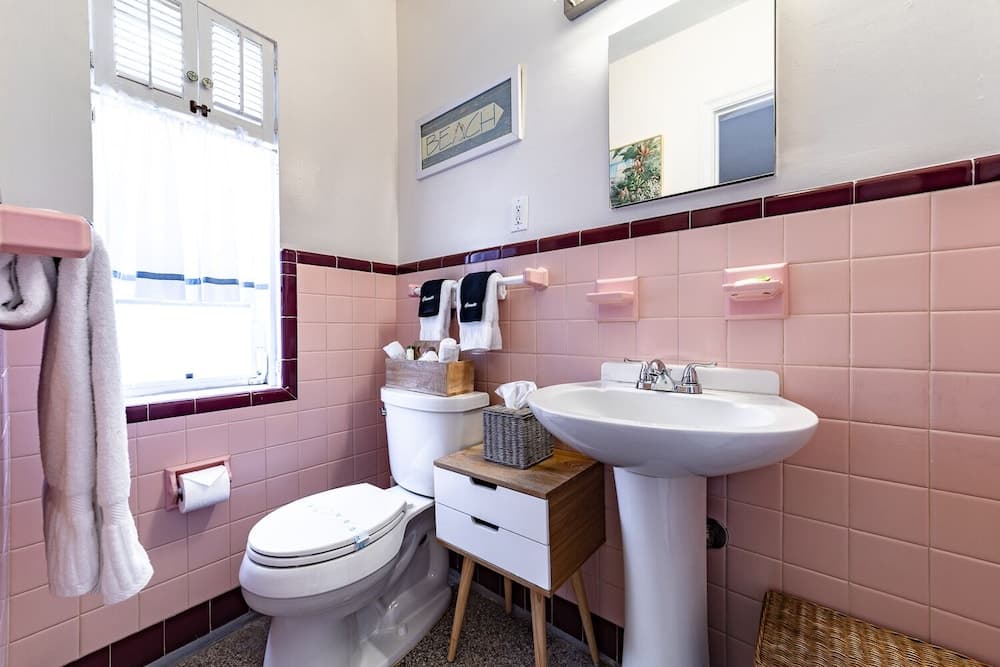 A charming vintage bathroom with pink tiled walls, white fixtures, and a cozy window view offers a nostalgic retreat for travelers seeking unique accommodations.