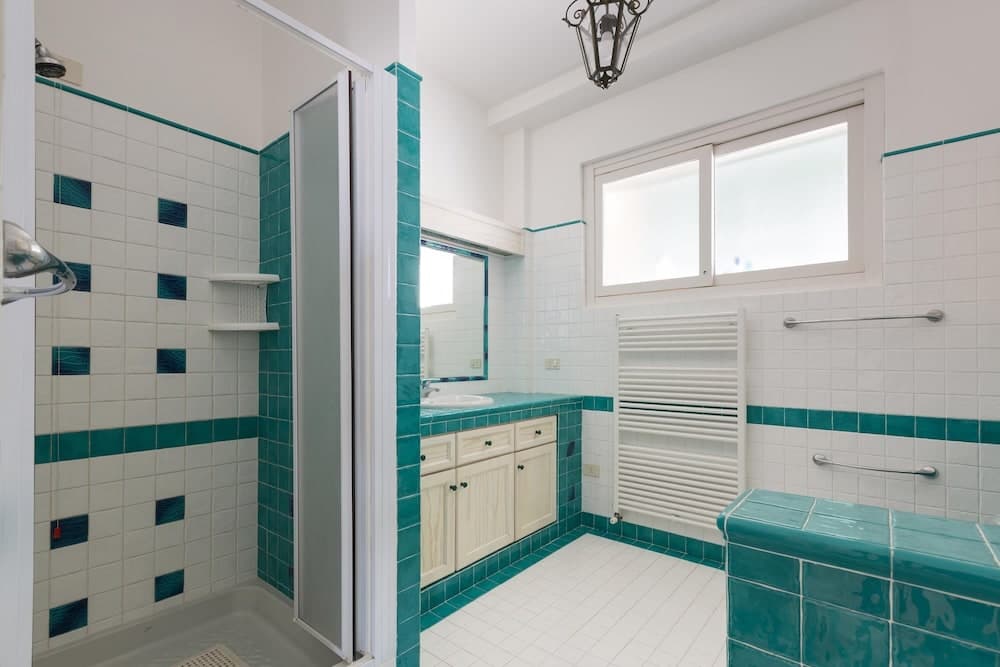 A bright, inviting bathroom features white tiles accented with teal stripes, a walk-in shower, a vanity with a mirror, and a window that lets in natural light, ideal for travelers seeking a clean and stylish accommodation space.