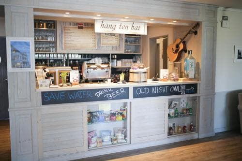 A cozy indoor bar with a rustic white counter, stocked shelves, quirky signs like "Save Water Drink Beer," and a guitar on display, evokes a relaxed, beachy vibe perfect for travelers seeking a laid-back spot to unwind.