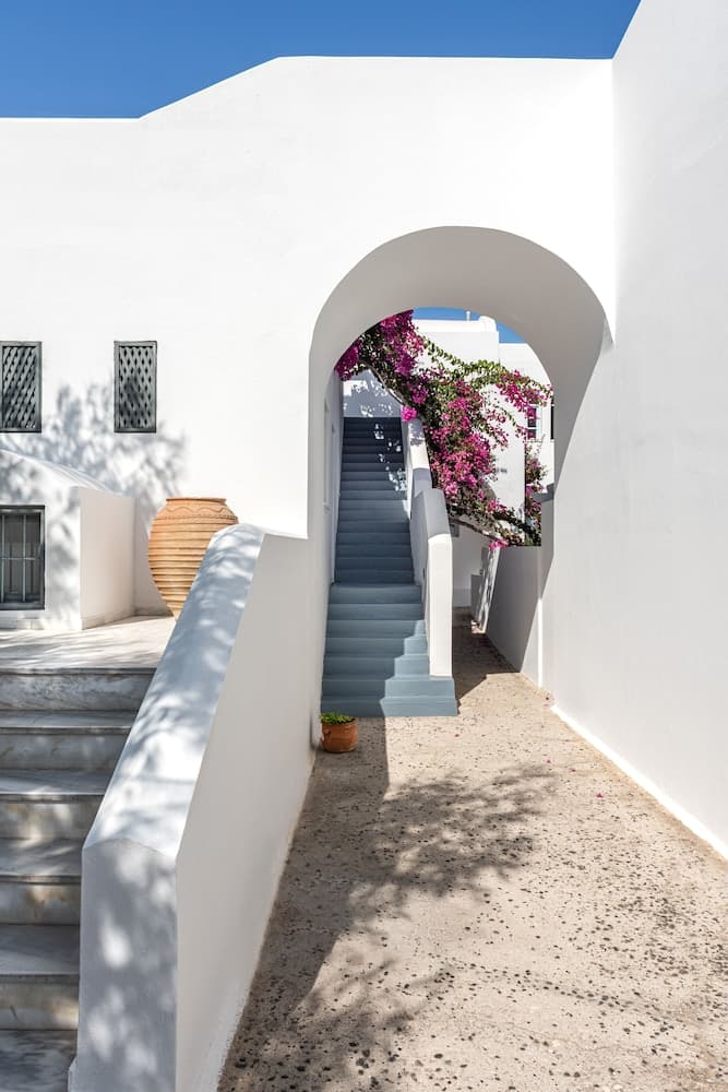 A serene Mediterranean courtyard features a white archway framing a staircase adorned with vibrant pink flowers, ideal for travelers seeking picturesque photo spots and tranquil retreats.