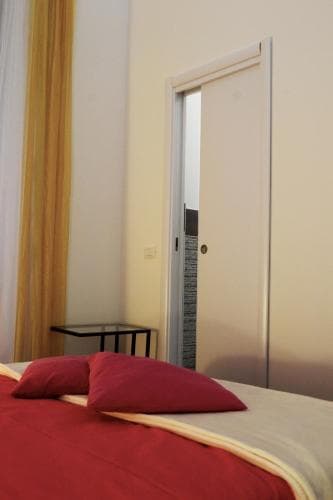 A cozy hotel room features a neatly made bed with red bedding, a small black side table, and a partially open door leading to a bathroom, ideal for travelers seeking comfort and simplicity.