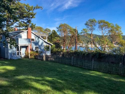 A serene coastal cottage with a wooden fence and lush green lawn overlooks a tranquil harbor, ideal for a peaceful seaside getaway.