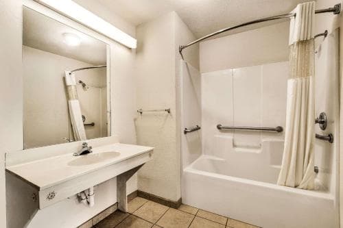 A clean, accessible hotel bathroom features a tiled floor, white fixtures, a large mirror, and a shower with grab bars, ideal for travelers seeking comfort and convenience.