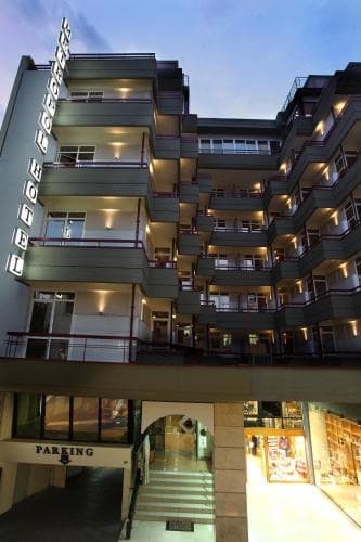 A warmly lit multi-story hotel with balconies overlooks a bustling shopping area at dusk, creating an inviting atmosphere ideal for travelers seeking accommodation near retail attractions.