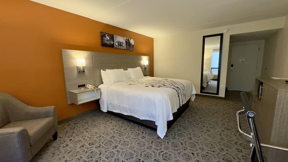 A cozy hotel room features a neatly made bed against an orange accent wall, modern furnishings including a chair and desk, and a full-length mirror, ideal for travelers seeking comfort and functionality during their stay.