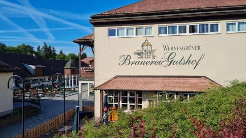 A charming outdoor seating area surrounded by lush greenery invites travelers to relax at the Wernesgrüner Brauerei Gutshof, an inviting brewery courtyard perfect for leisurely visits.