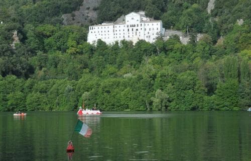 A serene lakeside scene features paddleboats adorned with Italian flags floating on calm waters, with a majestic white building nestled among lush green hills in the background, ideal for tranquil leisure trips.