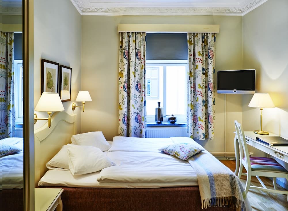 A cozy hotel room features a neatly made bed with floral curtains, warm lighting from bedside lamps, and a desk with a TV, ideal for travelers seeking comfort and functionality.