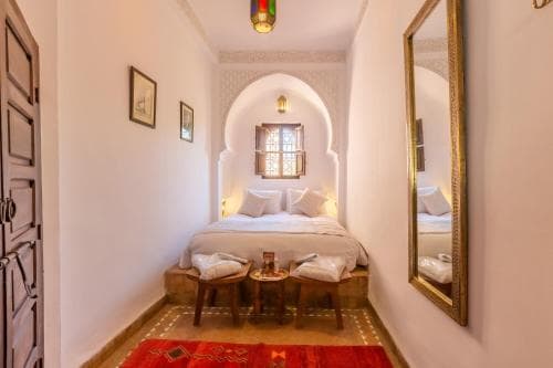 A cozy, warmly lit bedroom with Moroccan-inspired decor, featuring an arched window, ornate mirror, and patterned rug, ideal for travelers seeking a serene and culturally rich accommodation experience.