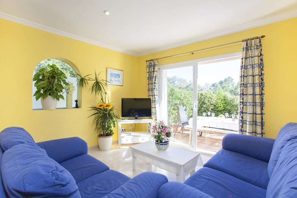 A cozy living room with vibrant yellow walls, blue sofas, potted plants, and a sliding glass door leading to a lush garden, ideal for travelers seeking a bright and inviting vacation rental space.