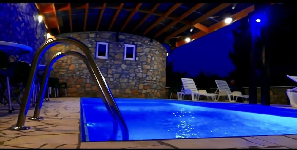 A serene evening at an outdoor pool area features a stone wall with small windows, lounge chairs, and a metal slide, illuminated by blue lighting under a wooden pergola, ideal for a relaxing vacation getaway.