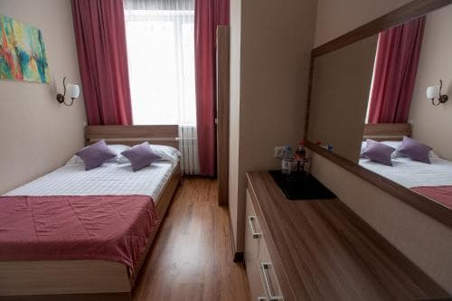 A cozy hotel room features a neatly made bed with red linens, wooden flooring, a large mirror, and vibrant artwork, creating a welcoming atmosphere ideal for travelers seeking comfort and relaxation.