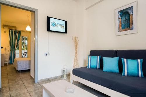 A cozy apartment interior features a navy blue sofa with turquoise pillows, a flat-screen TV, and an open doorway leading to a bedroom with green curtains, ideal for travelers seeking comfortable and modern accommodations.