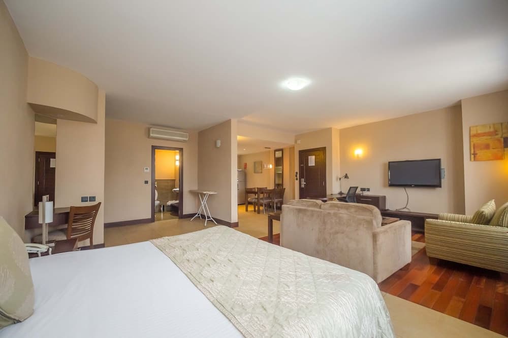 A cozy hotel suite features a neatly made bed, plush seating, a dining area, and warm lighting, ideal for travelers seeking comfort and relaxation during their stay.