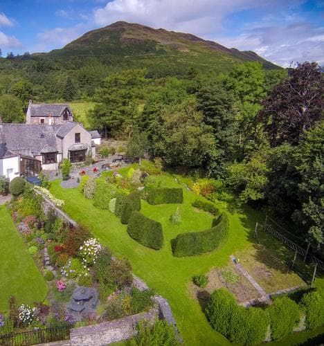 A serene countryside retreat features lush gardens with neatly trimmed hedges, vibrant flowerbeds, and a charming stone house nestled against a backdrop of rolling green hills, ideal for travelers seeking a peaceful escape in nature.