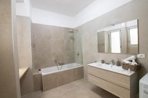 A serene modern bathroom with beige tiles, a bathtub, glass shower, wooden vanity, and large mirror, ideal for travelers seeking relaxation in a stylish accommodation.
