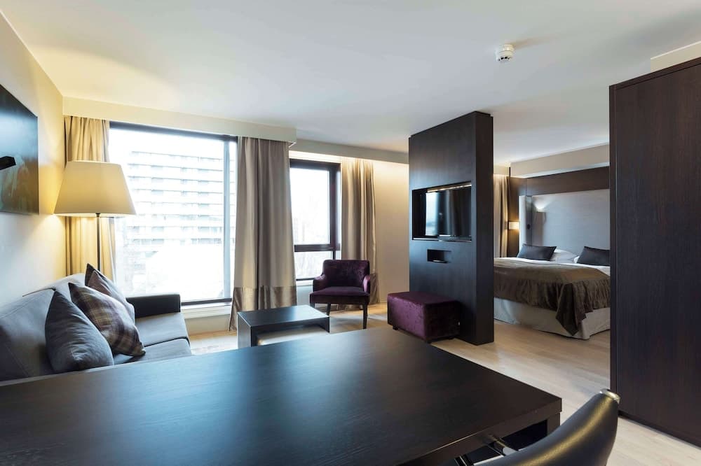 A cozy hotel suite features a modern living area with a sleek dining table, plush seating, and a bedroom separated by a stylish partition, ideal for travelers seeking comfort and functionality in an urban setting.