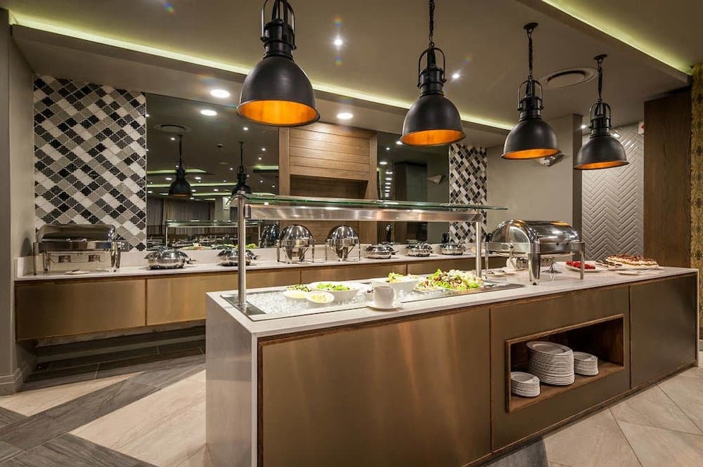 A modern buffet area in a hotel dining room features sleek metal counters, warm pendant lights, and an inviting array of food under glass domes, perfect for travelers seeking a comfortable and stylish dining experience.