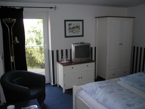 A cozy hotel room features a neatly made bed, a vintage TV on a white cabinet, a comfortable armchair, and a scenic view of greenery through the window, ideal for travelers seeking relaxation and comfort.