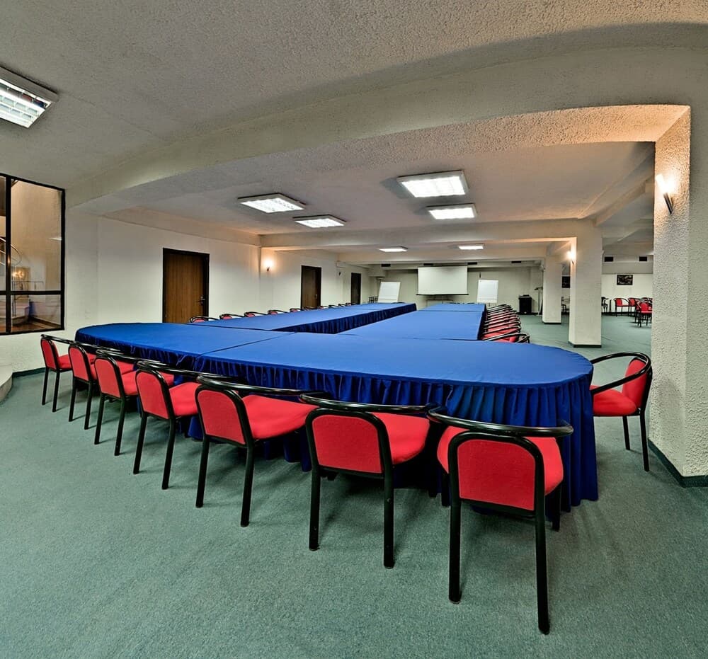 A spacious conference room with blue draped tables and red chairs under bright ceiling lights, ideal for professional meetings and seminars.