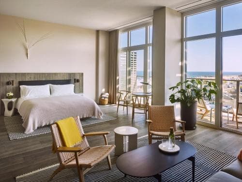 A cozy hotel room with a serene ocean view through large windows, featuring modern furnishings like a comfortable bed, stylish chairs, and a small dining area, ideal for travelers seeking relaxation and comfort by the seaside.