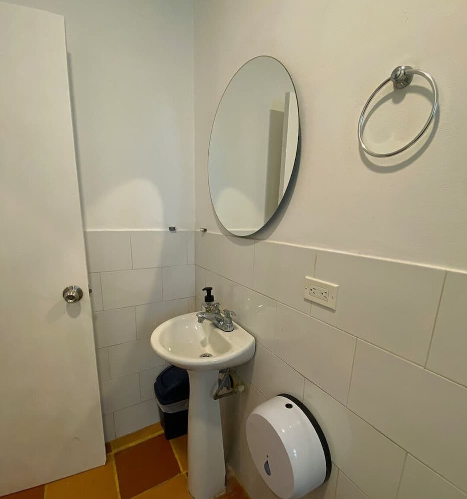 A compact bathroom features a white pedestal sink, round mirror, towel ring, and tiled walls, ideal for travelers seeking efficient space in accommodations.