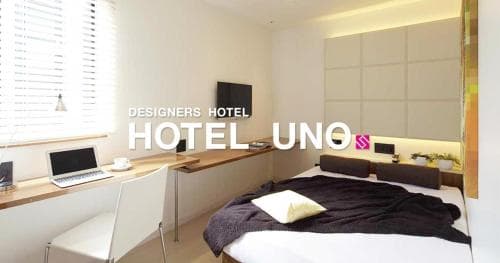 A modern designer hotel room features a sleek desk setup with a laptop by the window, a cozy bed with a minimalist design, and warm lighting, ideal for travelers seeking comfort and style in urban accommodations.