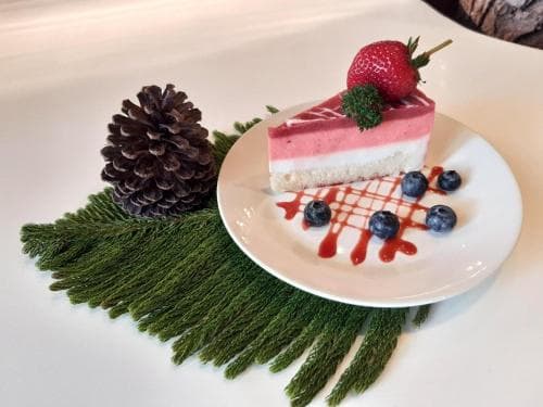 A beautifully plated slice of strawberry cheesecake garnished with blueberries and a strawberry sits atop a white plate, surrounded by festive greenery and a pinecone, creating a cozy holiday dessert scene perfect for seasonal celebrations.
