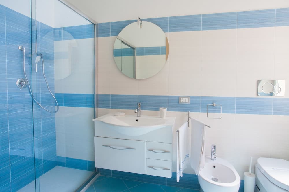 A serene bathroom with blue-tiled walls, a modern glass shower, a sleek white vanity, and a round mirror, ideal for travelers seeking a clean and stylish accommodation space.