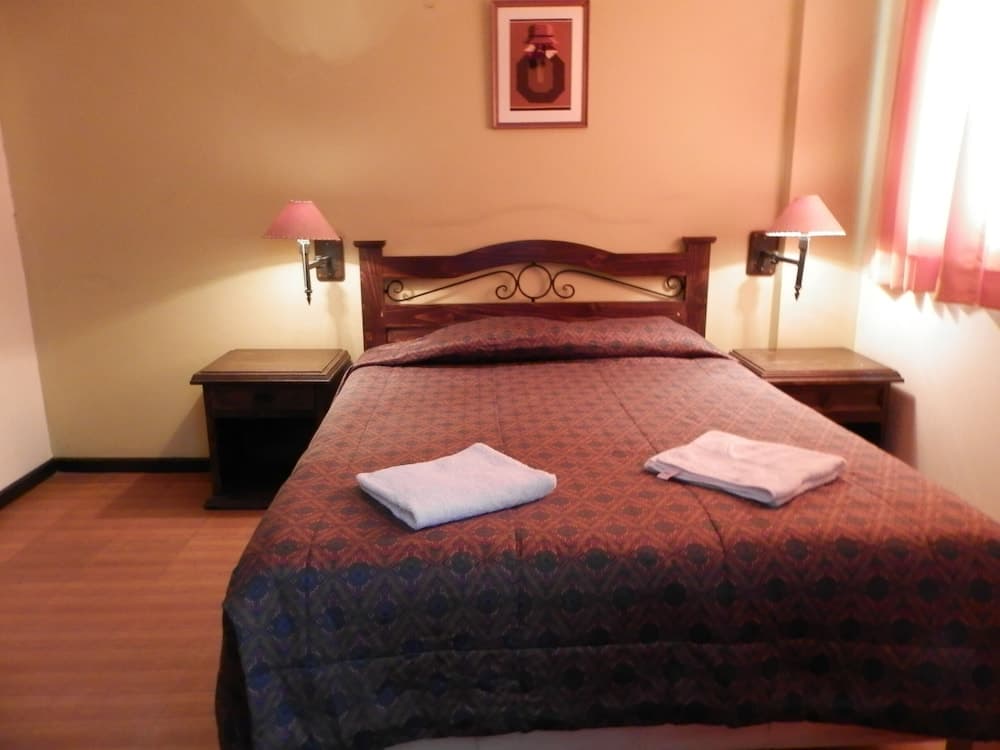 A cozy hotel room features a neatly made bed with patterned bedding, wooden furnishings, wall-mounted lamps, and soft natural light filtering through curtains, ideal for travelers seeking comfort and relaxation.
